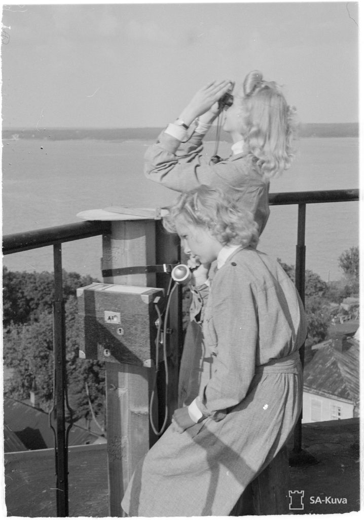 Två lottor står i kyrktornet i Sveaborgskyrka. Andra lottan spanar upp i luften med kikare, den andra sitter med telefonluren mot örat.