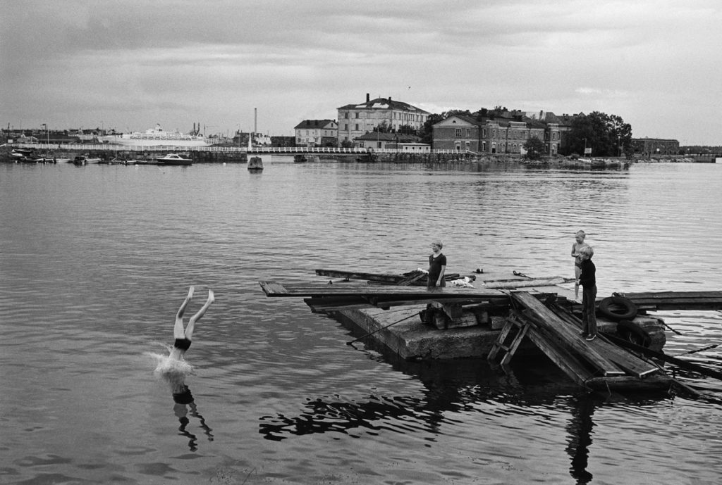 Barn på en brygga, en dyker i vattnet, i bakgrunden en ö med byggnader.