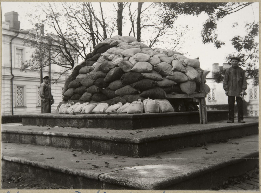 Två män står på varsinn sida av ett historiskt minnesmärke som är täckt med många sandsäckar.