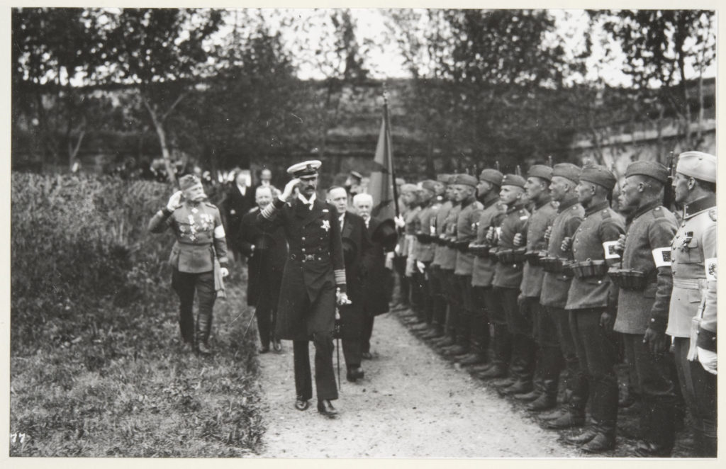 Historisk bild, uniformsklädd man gör honnör, soldater står i givakt till höger