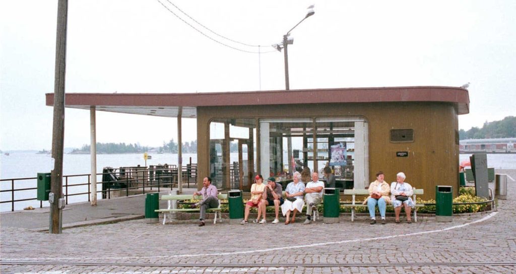 Sommaklädda människor sitter utanför en liten byggnad vid en kaj.