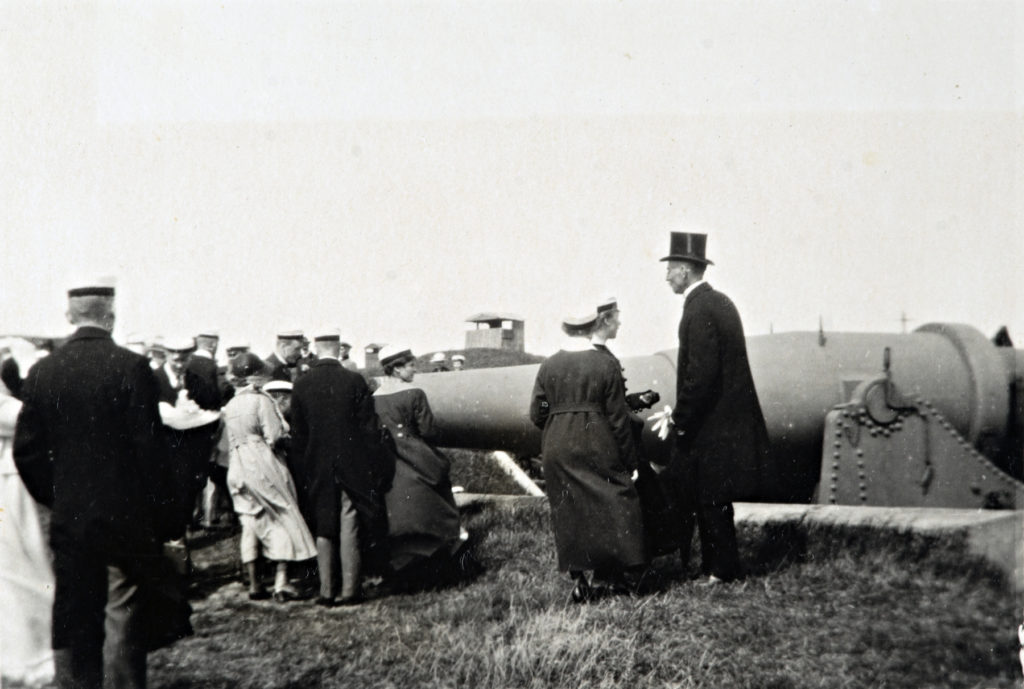 Män och kvinnor festklädda med hattar och studentmössor utomhus vid en kanon. Historisk bild
