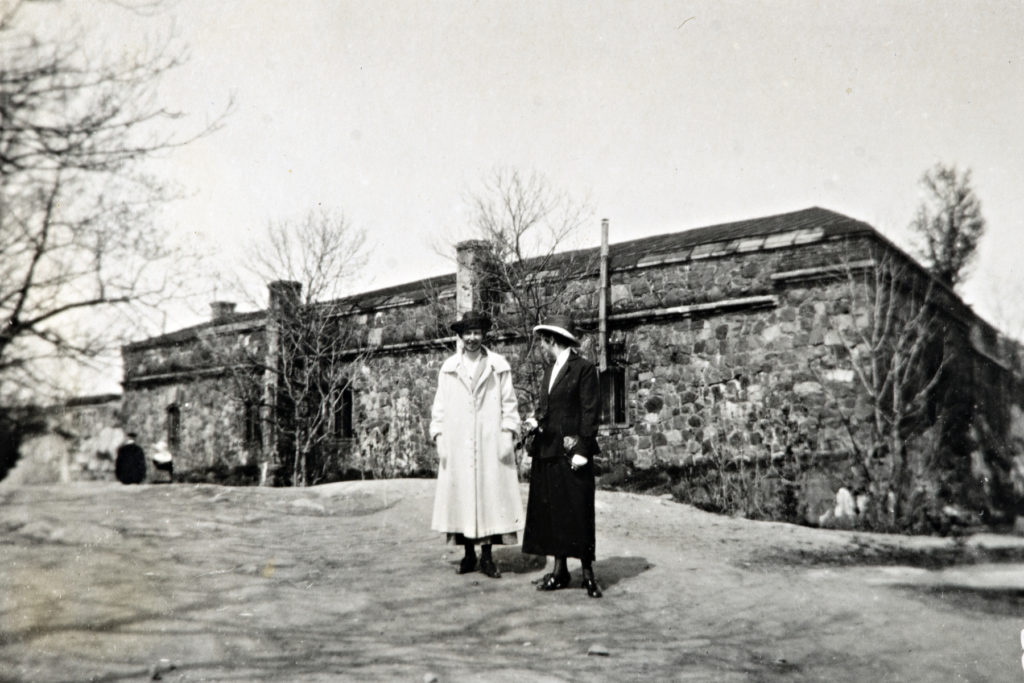 Två kvinnor står utomhus, i bakgrunden en del av ett fästningsverk. Historisk bild.