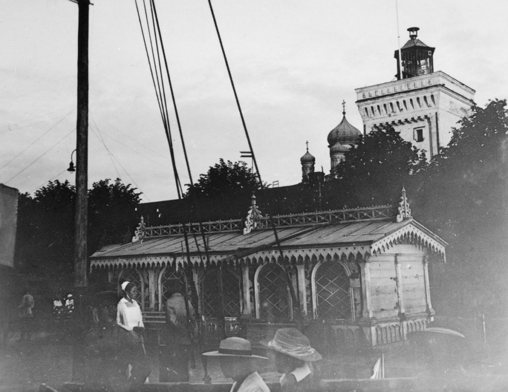 Läg träbyggnad med fönster med detaljrik arkitektur och utsirningar. I bakgrunden syns träd och byggnader, bl.a. kyrka med lökkupoler. I förbilden delar av ett skepp och några människor. Historisk bild.