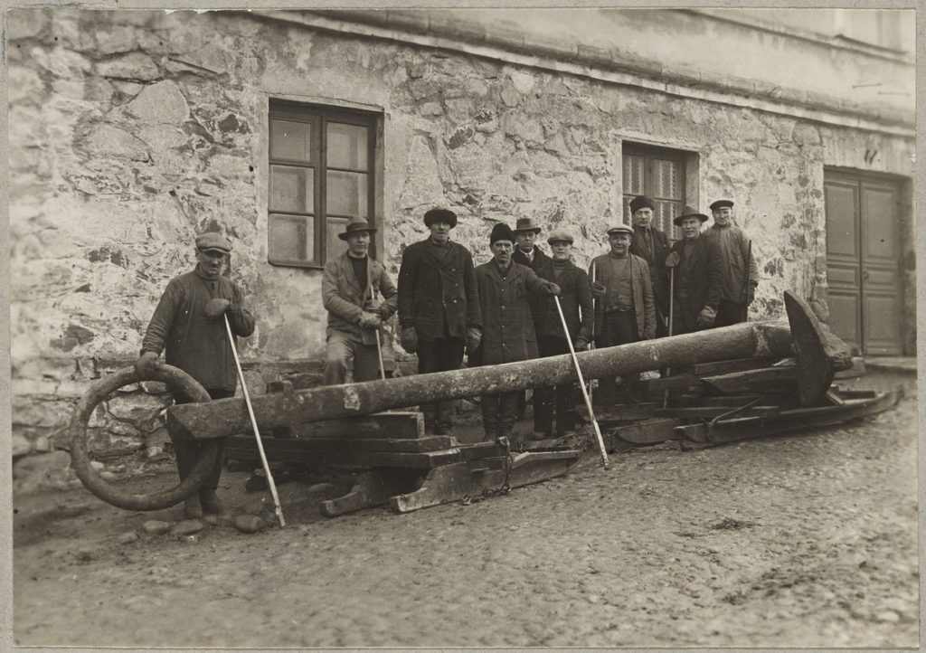 Tio män står bredvid ett enormt ankare utanför en gammal byggnad. Historisk bild.