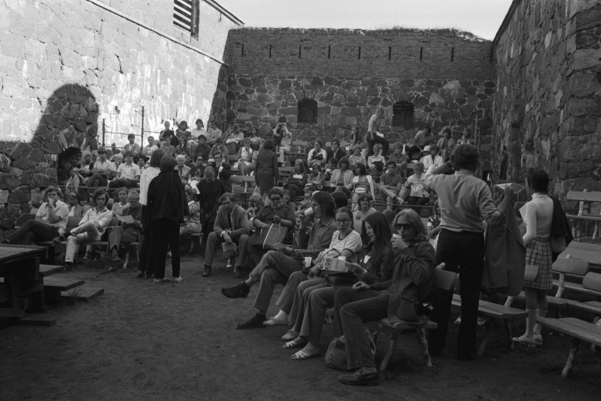 Många människor sitter utomhus på en gård omringad av gamla stenmurar. Svartvitt fotografi.