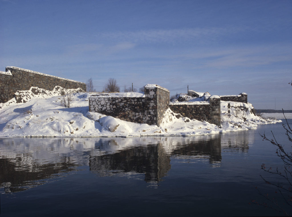 Gamla murar med snö på, vatten där isen inte ännu lagt sig. Blå himmel. Murarnas speglingar syns i vattnet. 