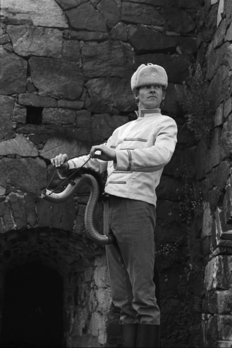 En man utklädd och med en låtsashäst står med en pälsmössa vid en gammal stenmur. Svartvitt fotografi.